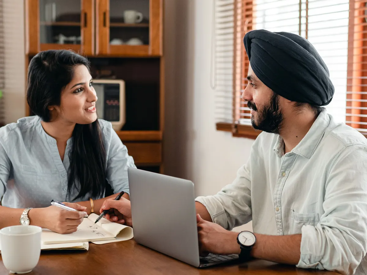 Two people talking over a notebook and a laptop at a wooden table